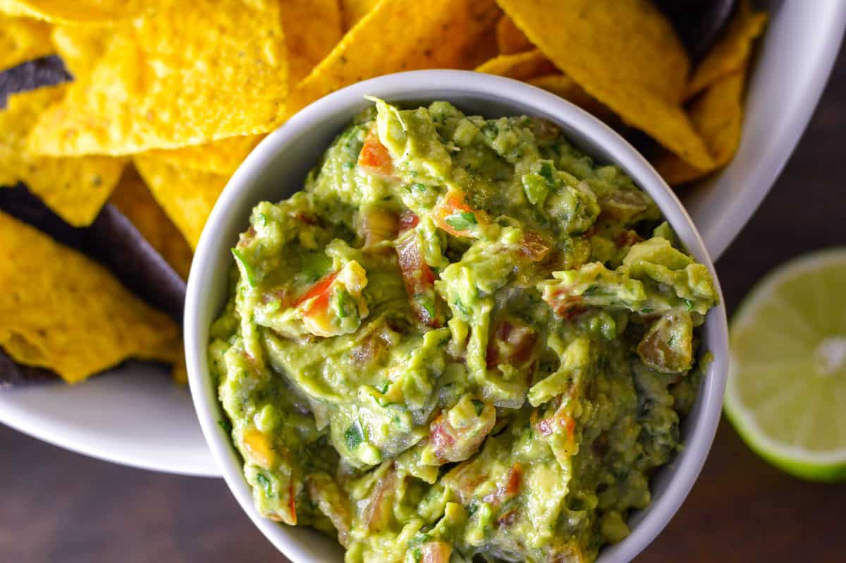 Simple Homemade Guacamole up close over top view in white bowl with yellow and blue chips surrounding with a half lime in background