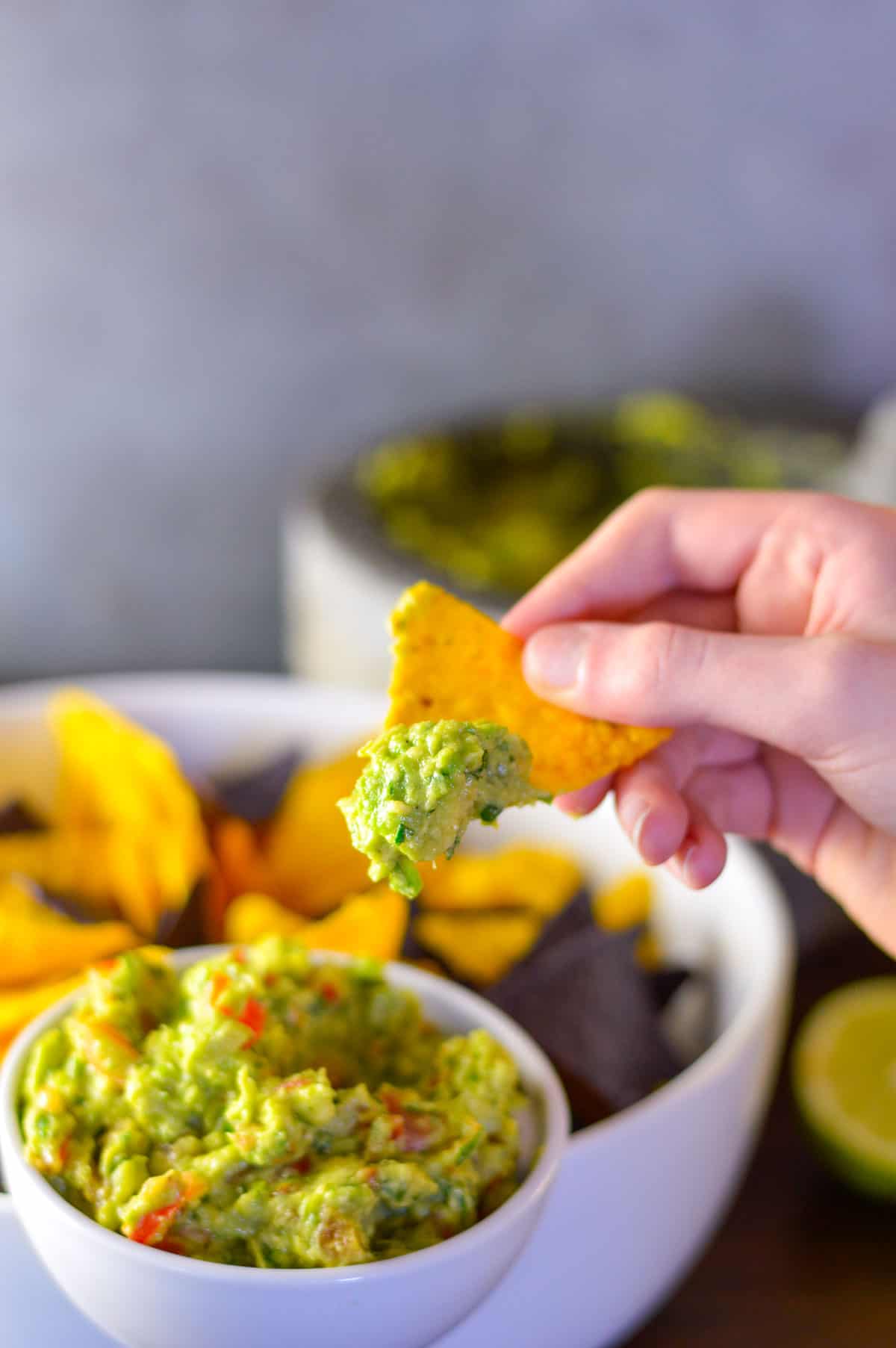 Chip being dipped in guacamole with bowl of chips and guac in background