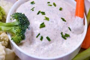 White bowl of Homemade Ranch Dip with carrot & broccoli dipped with more vegetables surrounding bowl & chives sprinkled on top