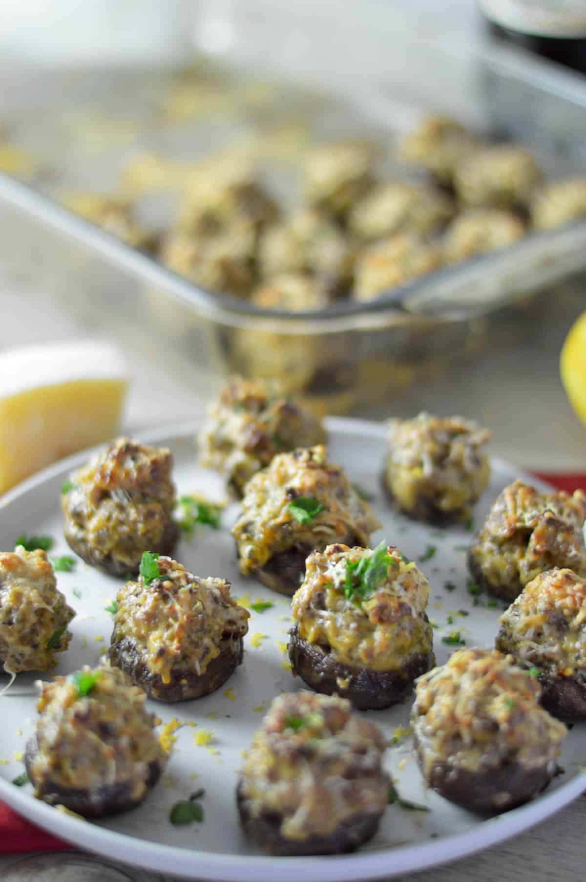 stuffed mushrooms on white plate with parsley and lemon zest sprinkled on top and parmesan and baking dish in the background