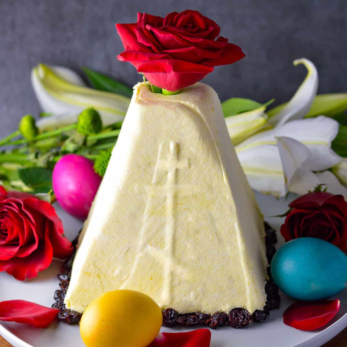 Orthodox Cross on side of paskha with a rose on top, surrounded by colored eggs, red rose and white flower in background