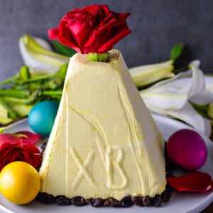 Paskha- Russian Crustless Cheesecake on white platter with colored eggs and red roses and red cranberries on the bottom.