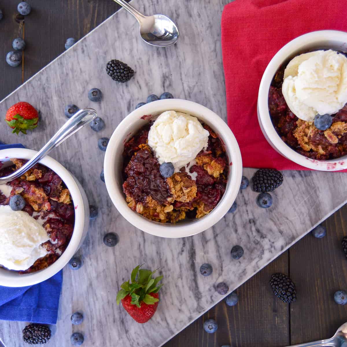 Slow Cooker Triple Berry Crisp dessert topped with vanilla ice cream in bowls with a couple spoons, berries on board and napkins