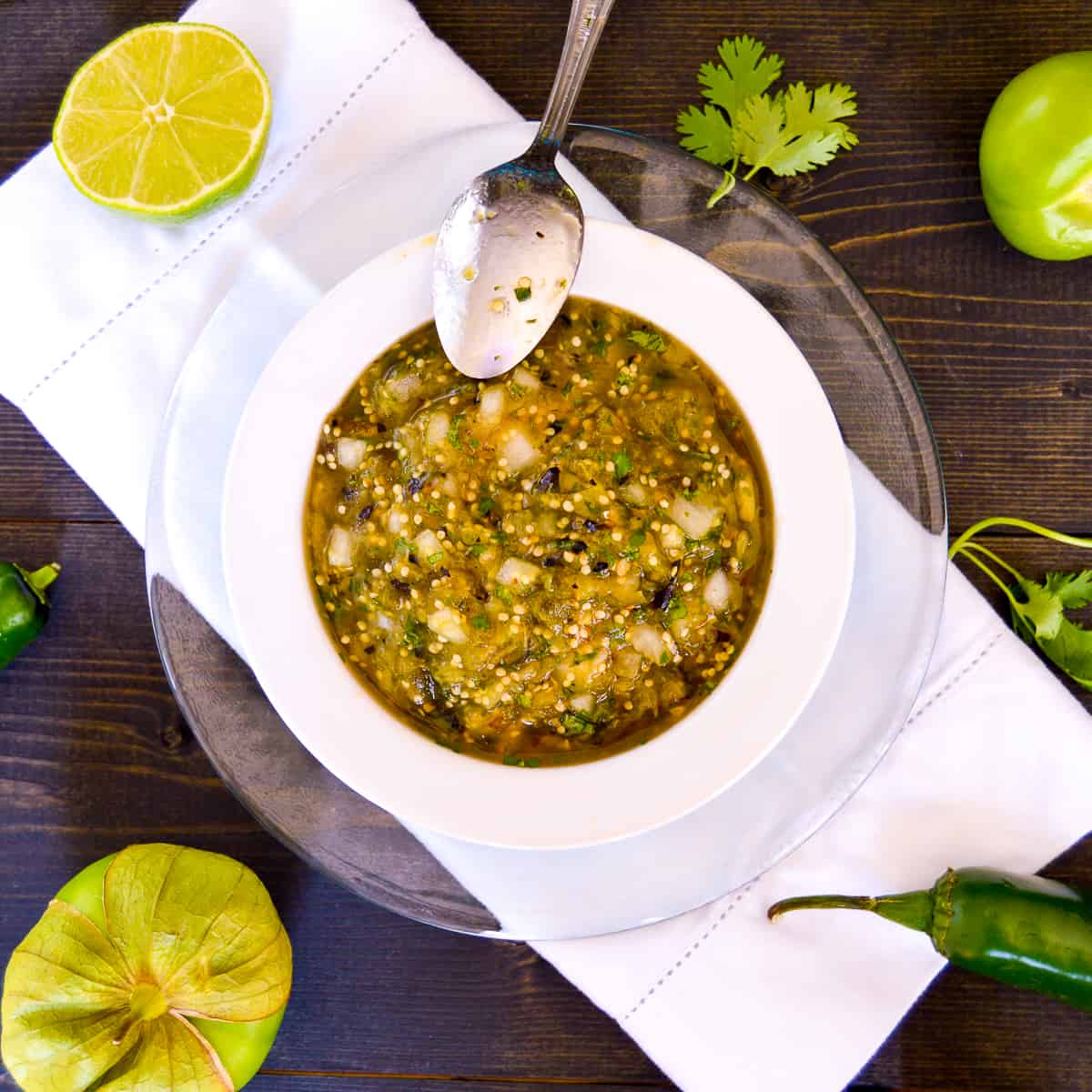 Over the top view of Tomatillo Green Salsa in white bowl, on glass plate with silver spoon and veggies on the side with cilantro