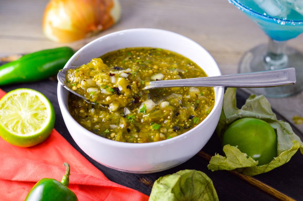 Spicy Salsa Verde in a white bowl with spoon resting on the side, on dark wood board with tomatillos, jalapeno and line. Onion and margarita glass in background