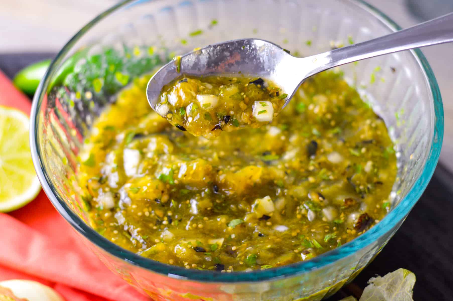 Glass bowl of spicy salsa verde with spoon of salsa on board with red napkin, lime and jalapeno in background