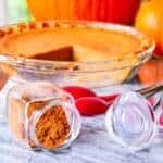 Pumpkin Pie Spice in clear spice jar overturned on marble with pie in background with pumpkins and measuring spoons