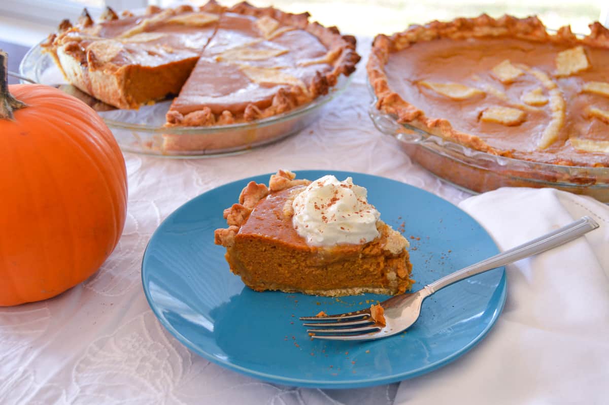 Pumpkin Pie slice with whipped cream and fork on blue plate with pumpkin and full pies in background