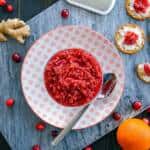 Raw Ginger Orange Cranberry Relish on red and white plate with a spoon on marble, fresh cranberries, orange and ginger beside with cream cheese and relish on crackers