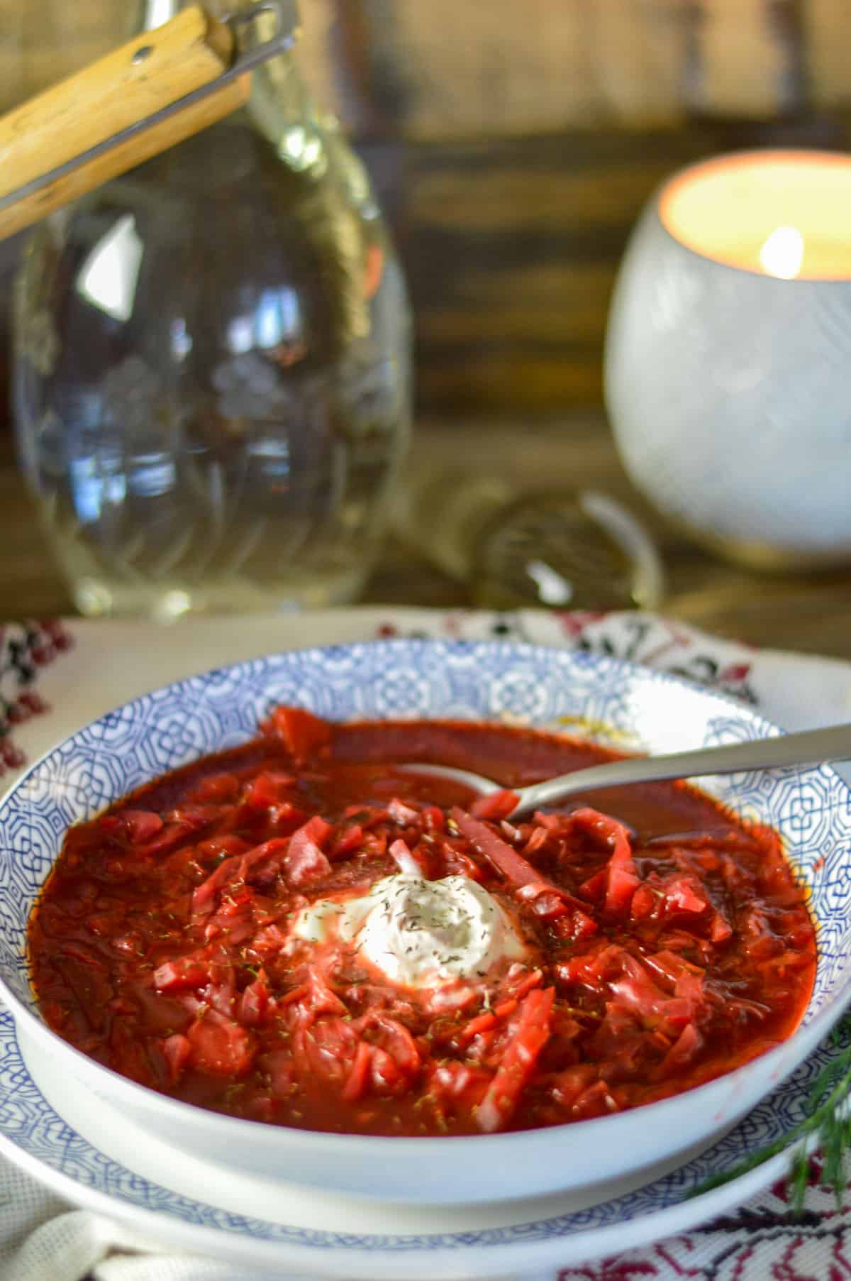 Red borscht in bowl with spoon, sour cream and dill. Glass bottle and candle in background with Ukrainian napkin