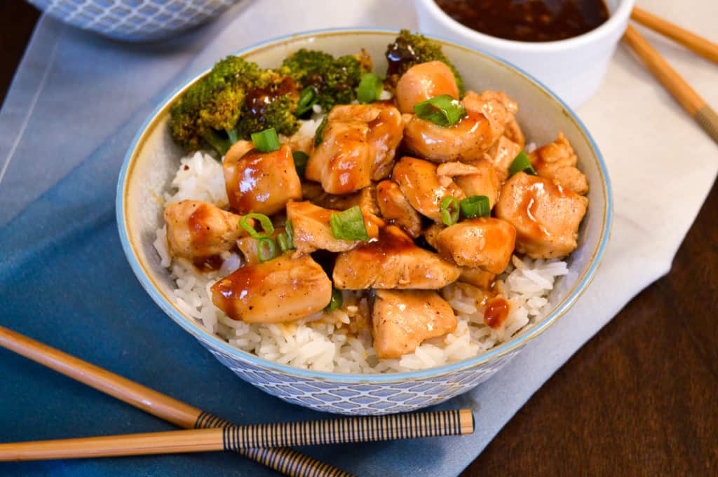 A bowl of steaming white rice topped with pieces of grilled chicken, broccoli florets, and garnished with chopped green onions. A small bowl of brown sauce and a pair of wooden chopsticks are placed beside the rice bowl.