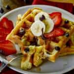 Waffle cut and forked on plate of waffles with banana, strawberries and syrup