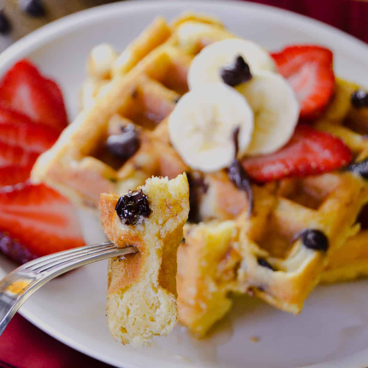 Buttermilk Chocolate Chip Waffles with up close shot of cut bite on fork with bananas and strawberries and maple syrup