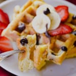Up close shot of Buttermilk Chocolate Chip Waffles cut on fork with waffles in background with strawberries, bananas and maple syrup on top