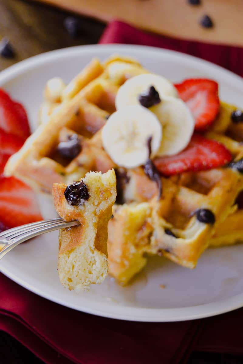 Up close shot of Buttermilk Chocolate Chip Waffles cut on fork with waffles in background with strawberries, bananas and maple syrup on top