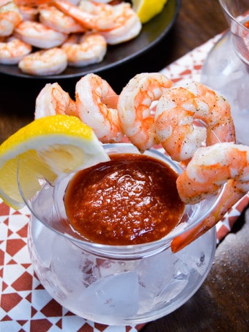 Roasted Shrimp Cocktail in Martini glass with cocktail sauce and lemon wedge with more shrimp in background, on red and white napkin