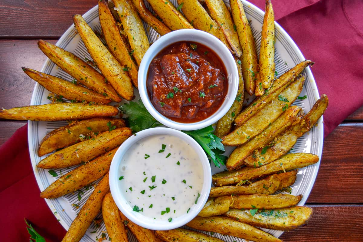 Plate of air fyer potato wedges on plate with parsley and dips, parsley sprinkled on top, shot from above