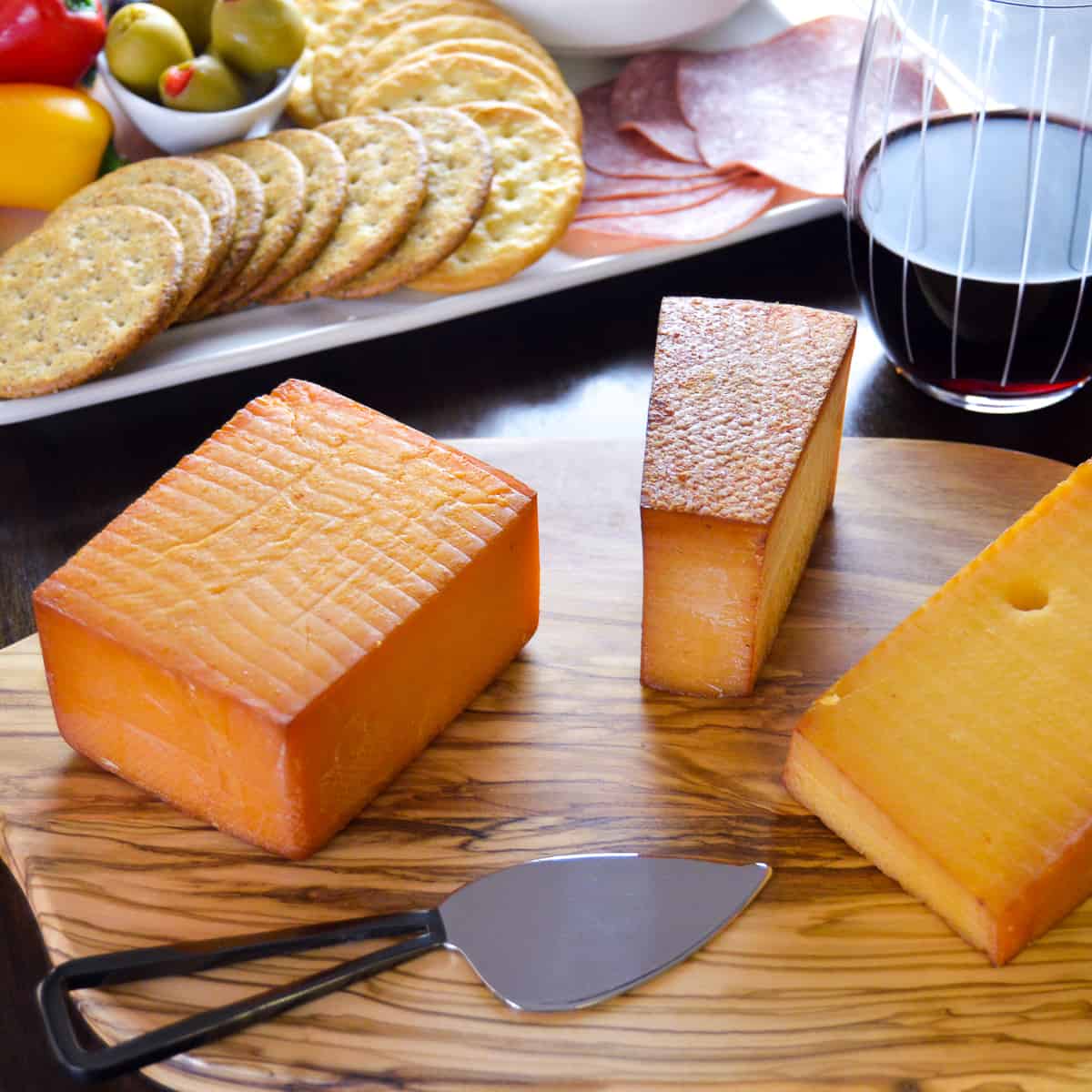 Cold Smoked Blocks of Cheese. Cheddar, Gouda, Gruyere & plate of crackers, olives, peppers, grapes, salami in background with glass of red wine