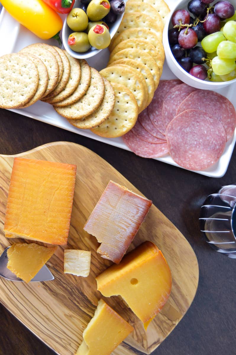 Smoked cheese on wooden cutting board with appetizer plate with crackers, grapes, crackers, olives and peppers in background with glass of red wine