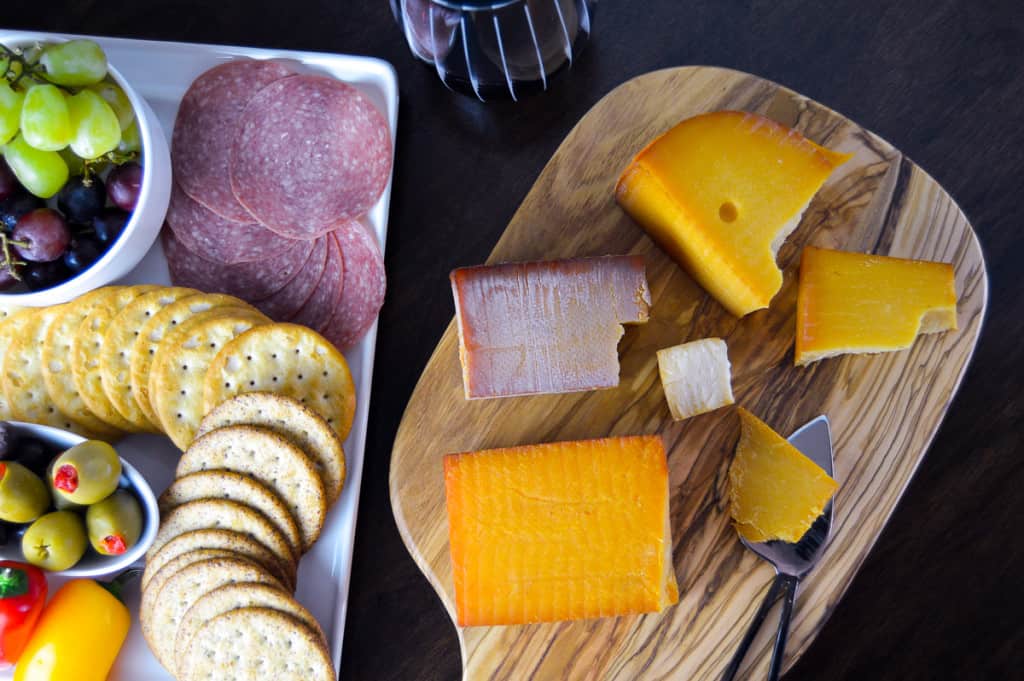 Over-top view of smoked cheeses on wooden cutting board, glass of red wine, and plate of salami, crackers and veggies, grapes