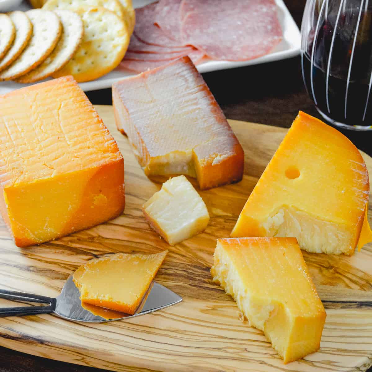Cold Smoked Cheese with broken off pieces, a slice of smoked cheddar with a glass of red wine, crackers and salami in background