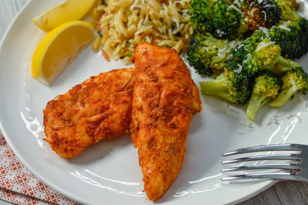 Chicken Tenders on white plate with fork, lemon wedges, orzo and cooked air fryer broccoli