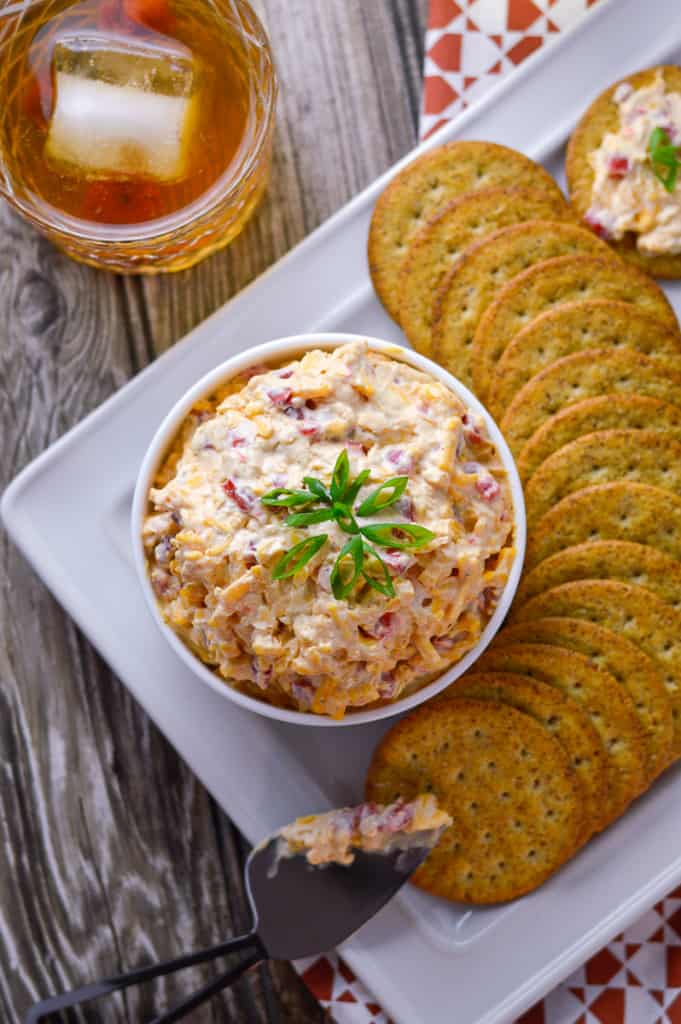Pimento cheese spread in white bowl, surrounded by crackers with a knife and bourbon glass on the side