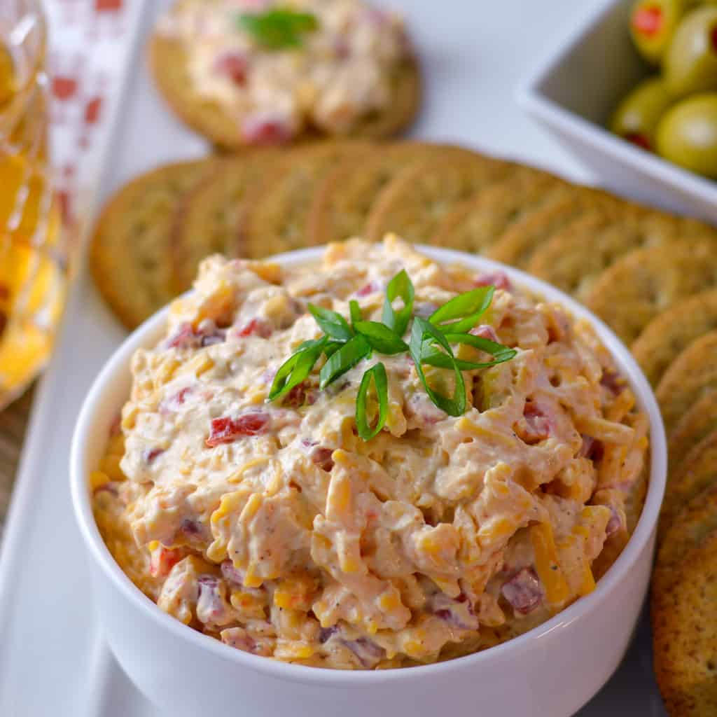 super close of view of pimento cheese spread with green onions on top with crackers, olives and glass in background