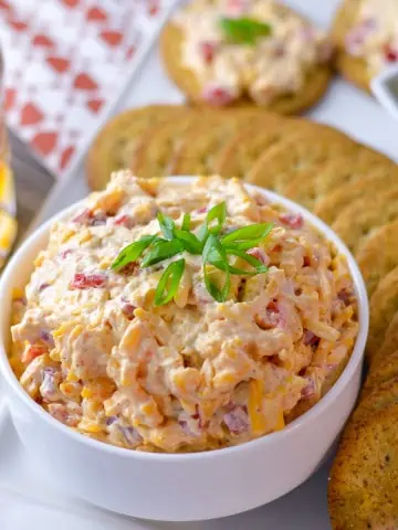 Featured image of Southern Pimento Cheese Spread in white bowl with green onion, surrounded by crackers, some green olives, topped crackers with spread and a glass of bourbon