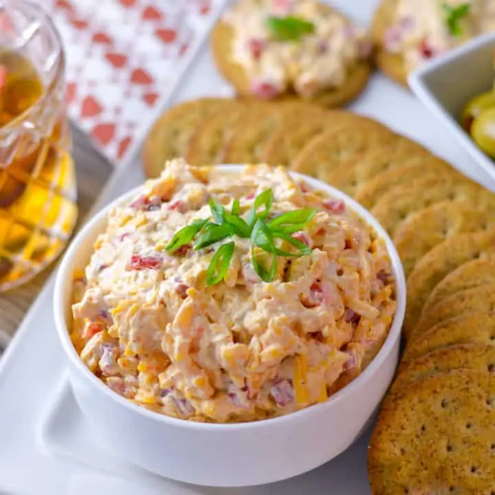 Featured image of Southern Pimento Cheese Spread in white bowl with green onion, surrounded by crackers, some green olives, topped crackers with spread and a glass of bourbon