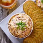 Pimento cheese spread in white bowl, surrounded by crackers with a knife and bourbon glass on the side
