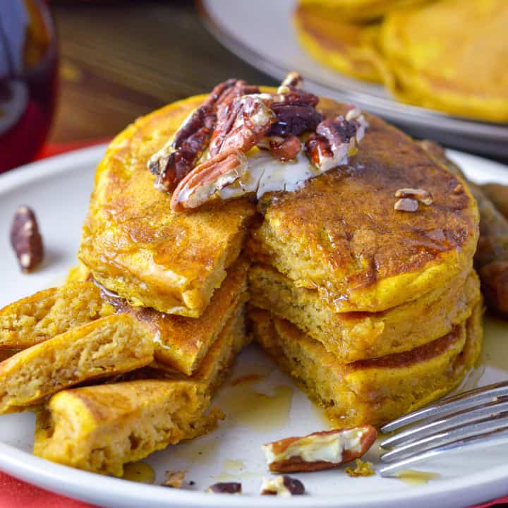 3 Fluffy Pumpkin pancakes with cut out of side with pecans, syrup and syrup bottle and plate of pancakes in background