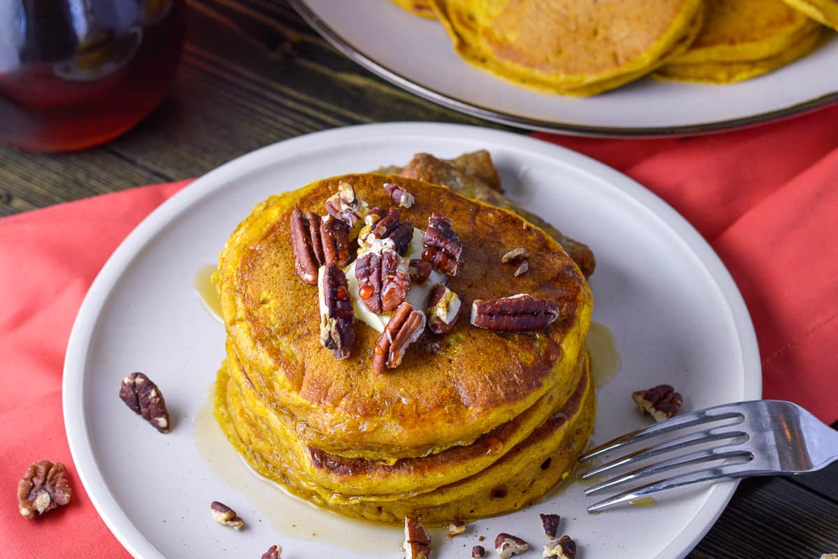 3/4 view of 3 stack of pumpkin pancakes with pecans and syrup on plate with sausage