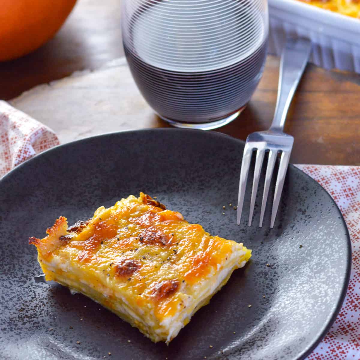 Square of au gratin dauphinoise potatoes on black plate with pumpkin, red wine and casserole dish with au gratin in background