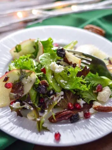 Holiday Green Salad with Apple and pomegranate on white plate with green napkin and white lights in background