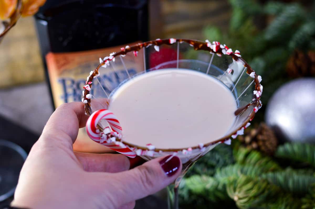 Hand holding glass of Bourbon Chocolate Martini with bourbon cream in background with evergreen, ornament and staves