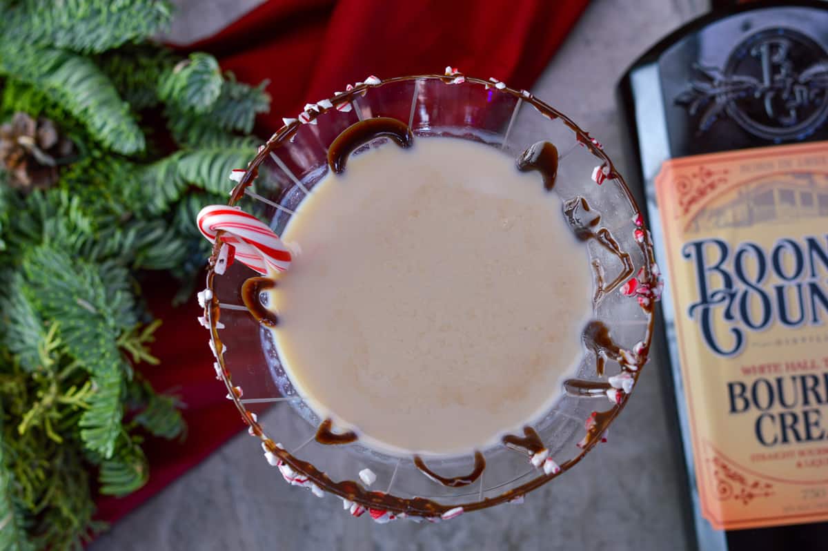 Close up over the top of Bourbon cream martini with candy cane pieces on rim with syrup and mini candy cane and evergreen in background