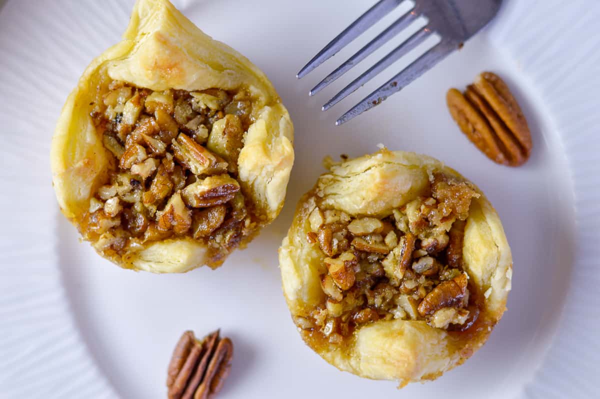 Close up of 2 pastry puffs with maple pecan pie filling on white plate with fork and pecans on the side