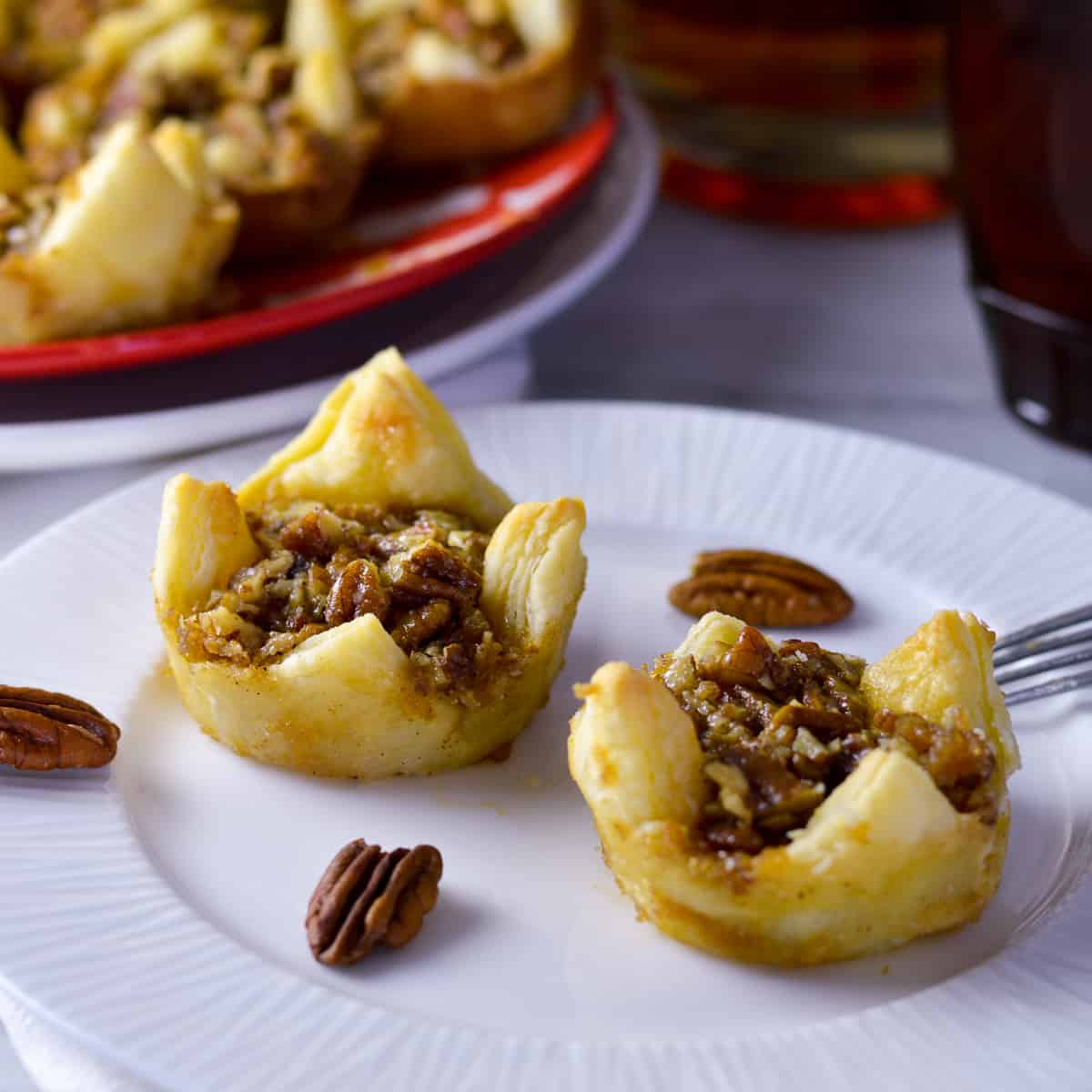 Side view of pecan puffs on white plate with fork and pecans and more puffs on red and white plate in background with maple syrup and bourbon