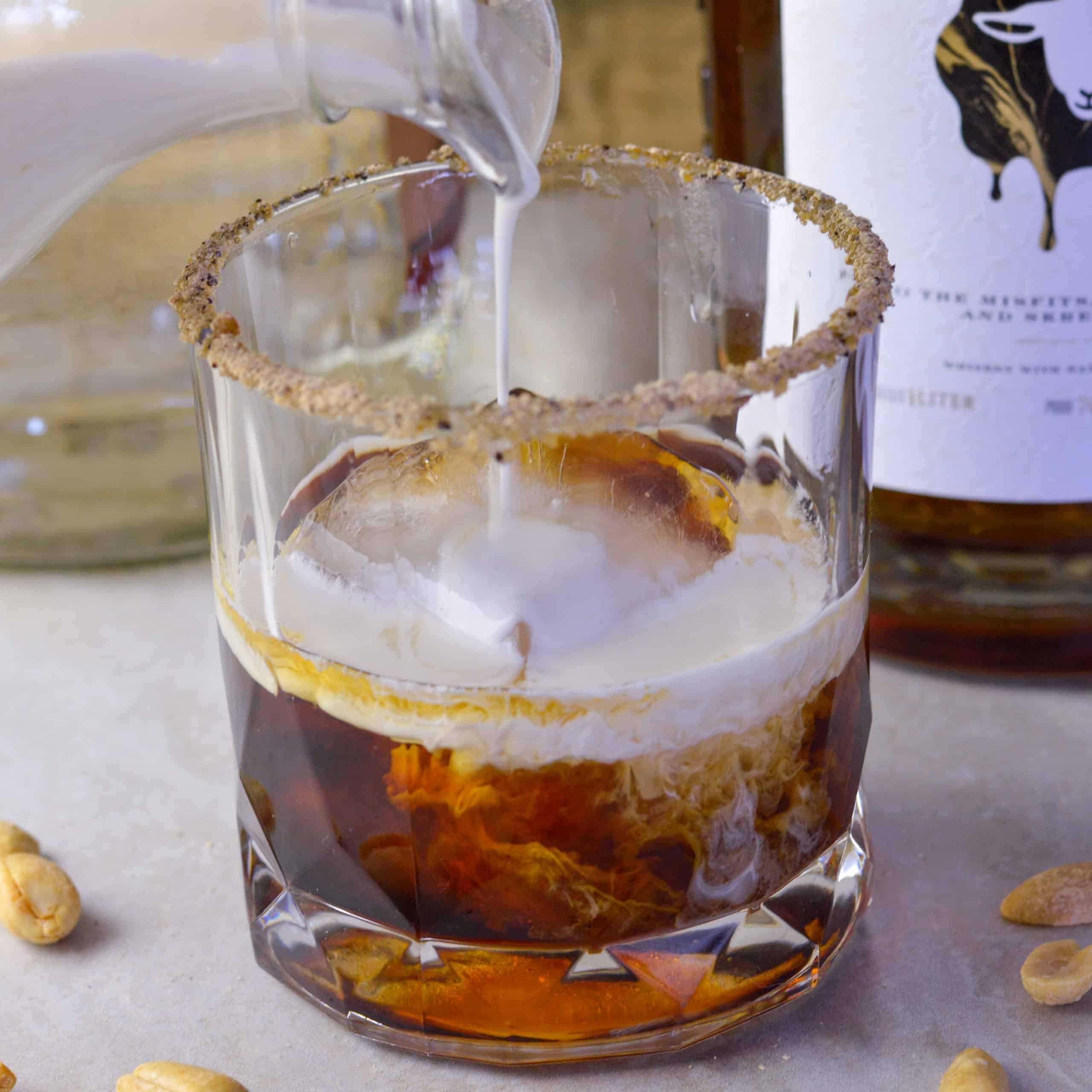 Adding a splash of heavy cream to a Skrewball White Russian Cocktail with Skrewball bottle in background and peanuts on counter