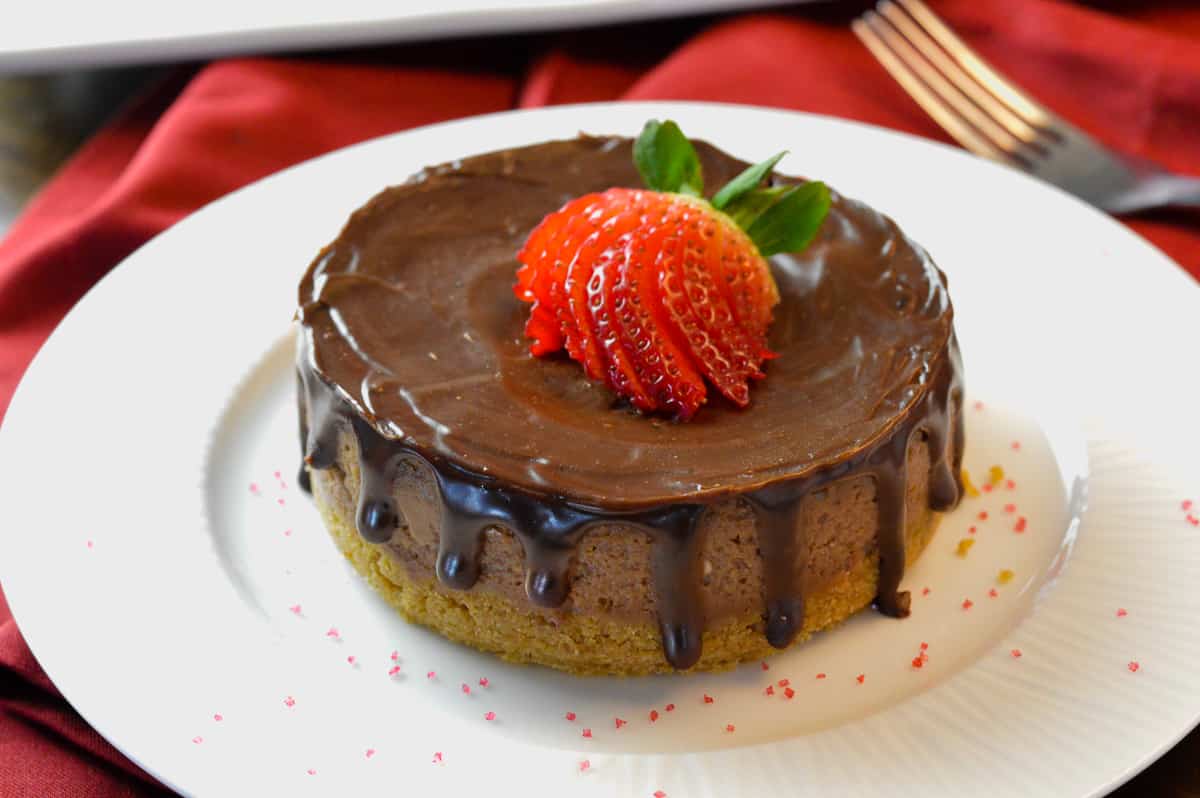 Nutella cheesecake with chocolate ganache drips and splayed strawberry on top on white plate with sprinkles on red napkin with silver fork