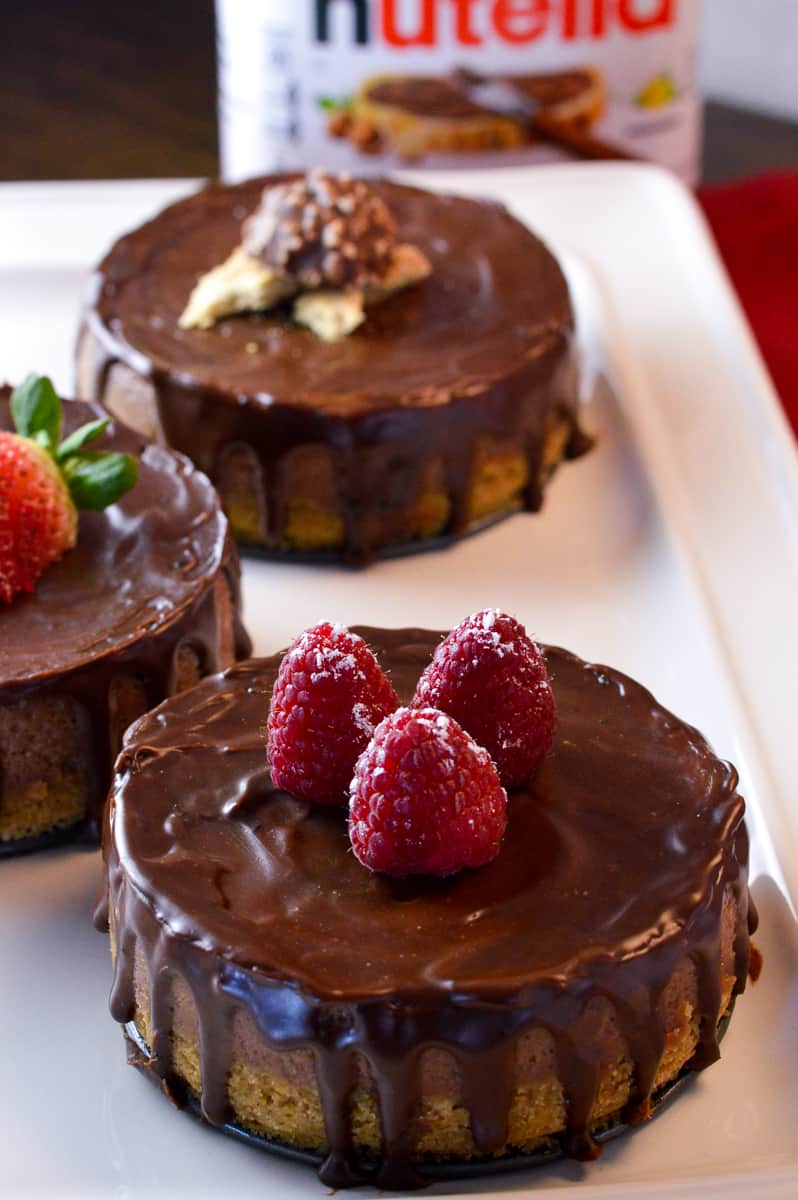 3 Nutella cheesecake minis on white platter with raspberries, strawberry and ferrer rocher with jar of nutella in the background