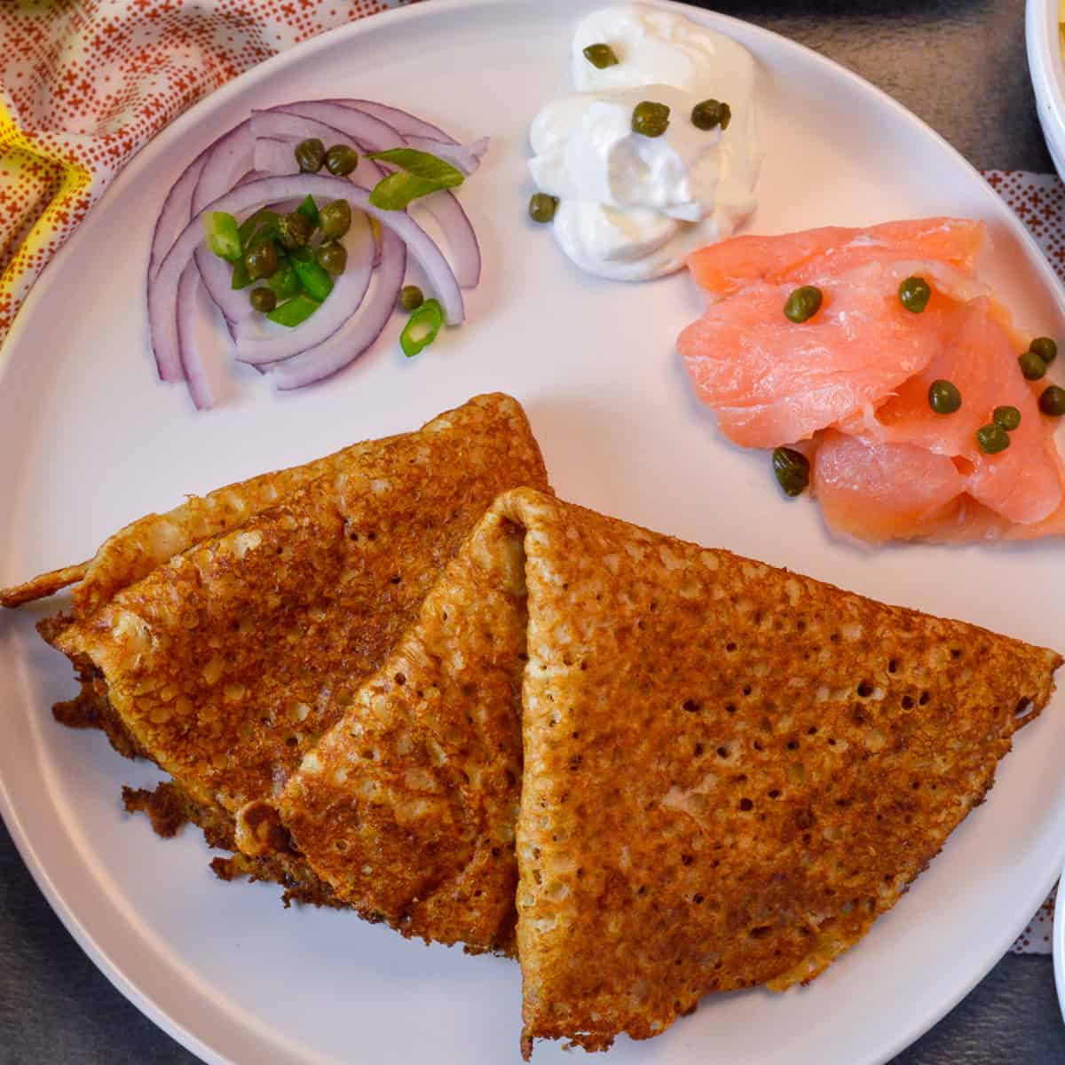 2 blini folded into fourths on plate with red and green onion, sour cream and smoked salmon with capers on red and white napkin