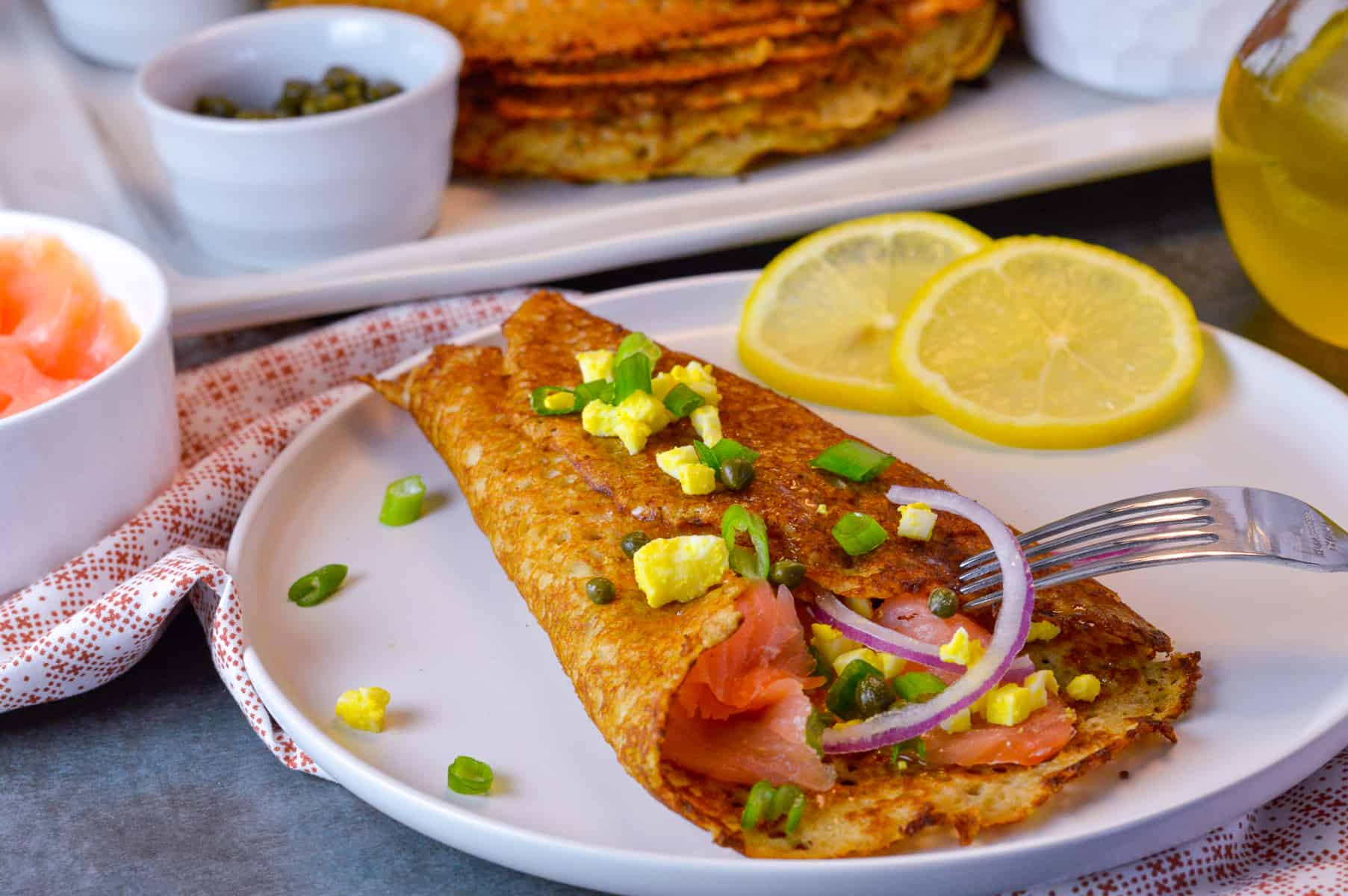 blin folded up on plate, with fillings and some hard boiled eggs, green onion and red onion on top with lemon on side and smoked salmon, capers and blini stack with melted butter in background