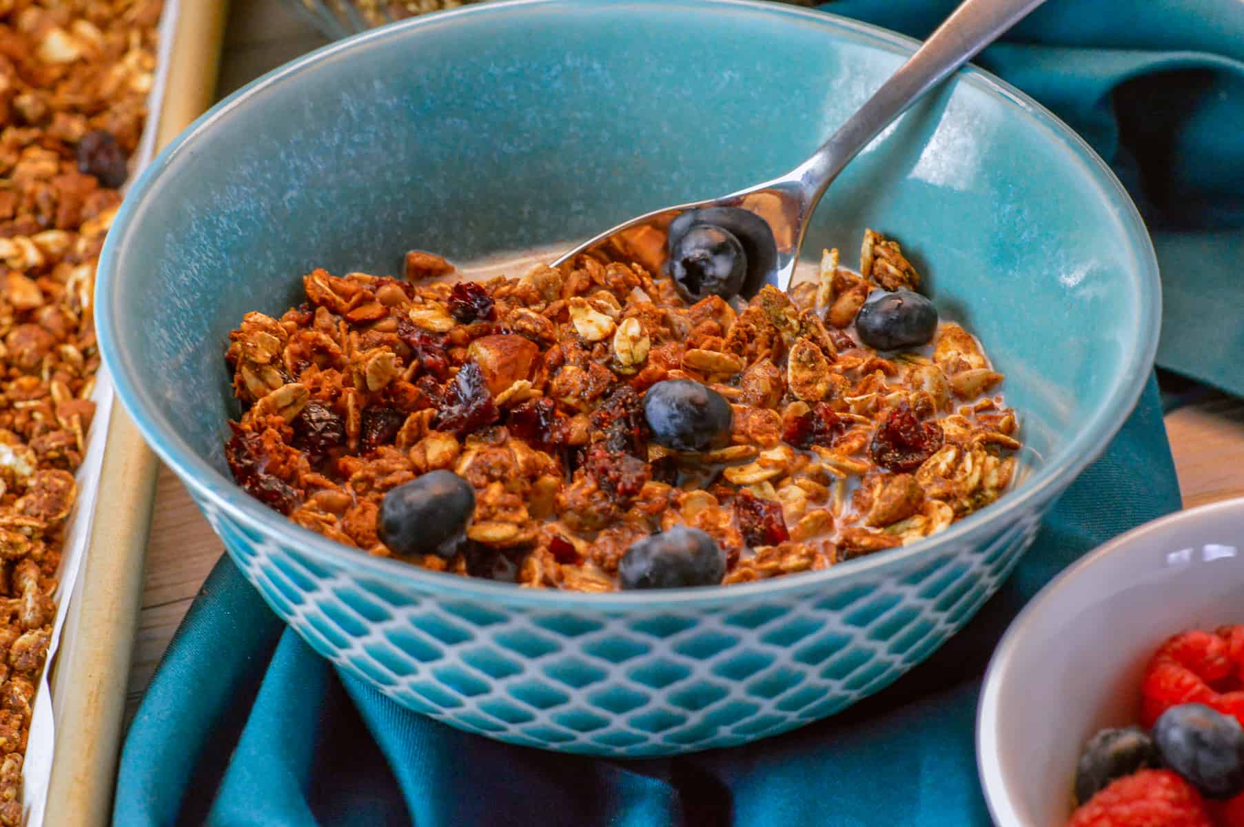 bowl of granola high protein cereal in teal bowl with berries and more berries in a white bowl on the side on a dark teal napkin