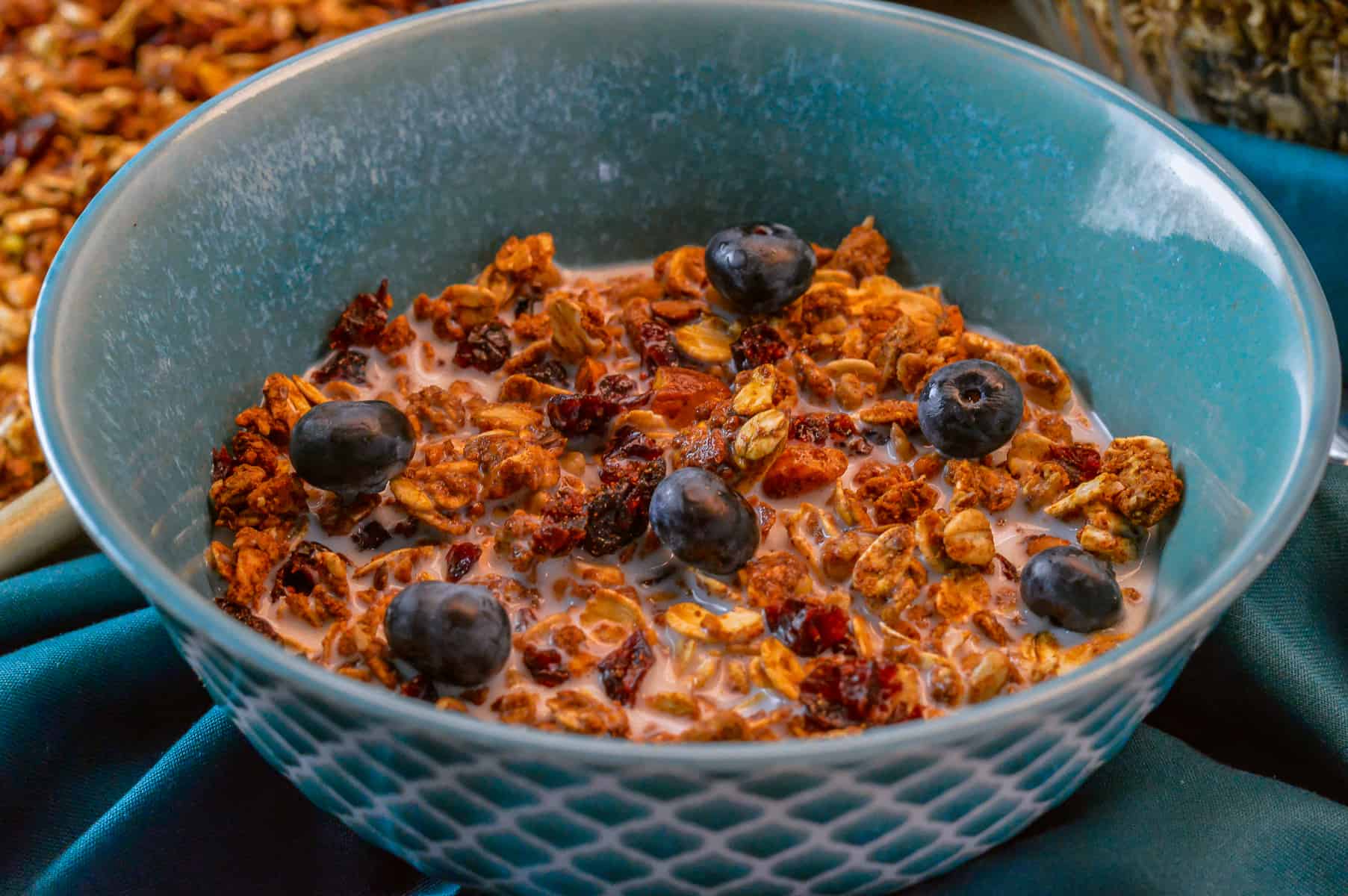 bowl of granola with milk and blueberries in teal cereal bowl on dark teal napkin with granola in background