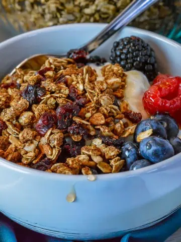 High protein granola in white bowl with yogurt, honey dripping of side with blackberries, raspberries, blueberries and big bowl of granola in background