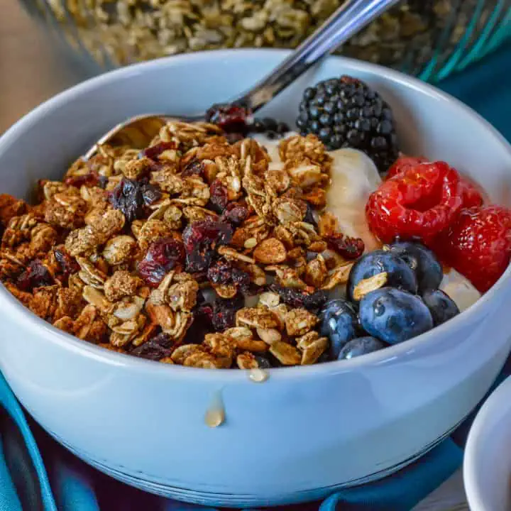 High protein granola in white bowl with yogurt, honey dripping of side with blackberries, raspberries, blueberries and big bowl of granola in background