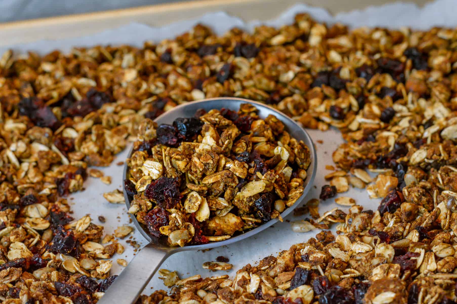 Granola with almonds and dried cranberries close up on silver spoon with more granola on sheet pan with parchment paper