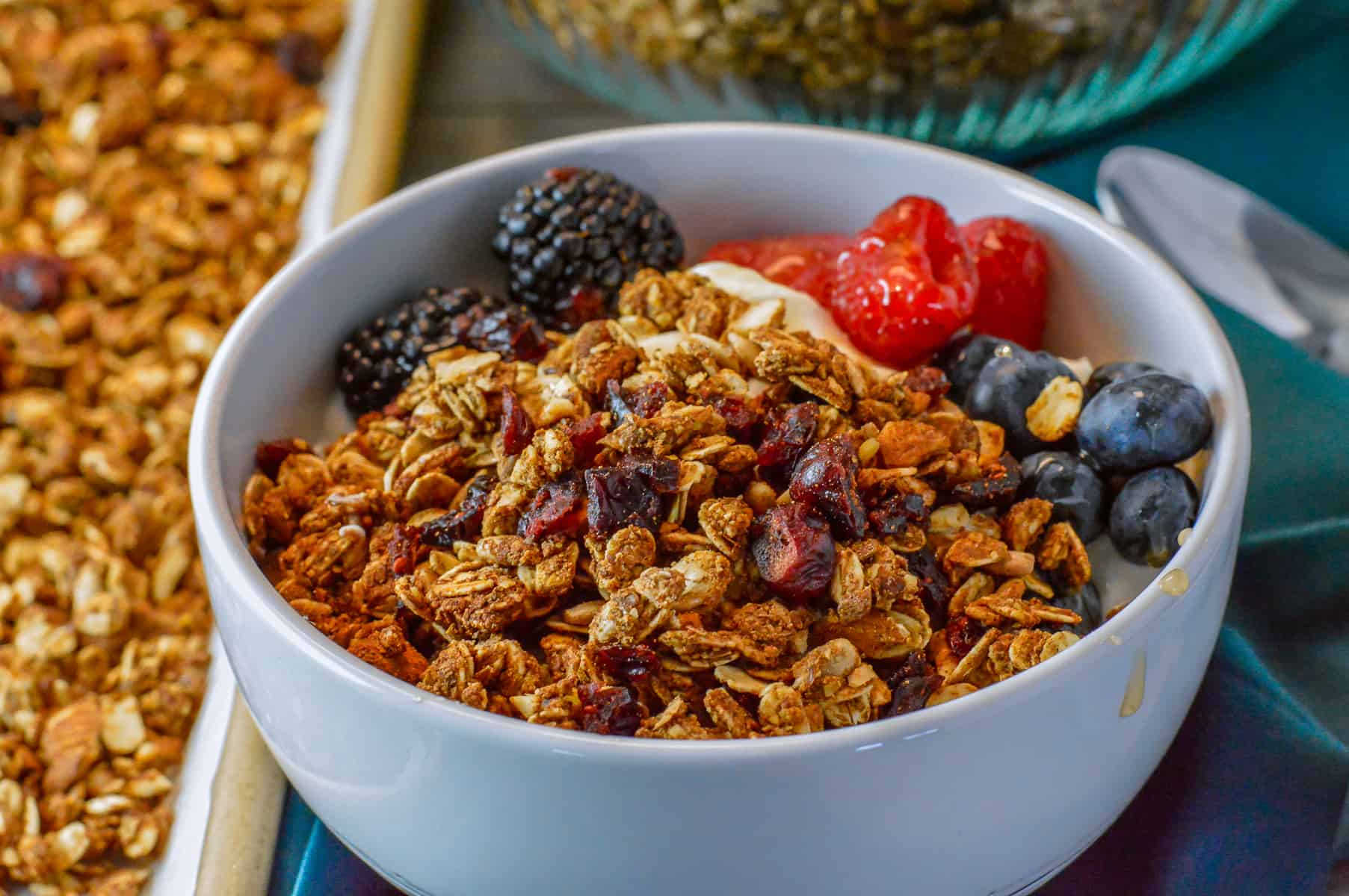granola in white bowl with yogurt, honey, blackberries, raspberries, and blueberries with baking sheet with granola on side and bowl in background with silver spoon on the side